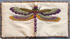 an embroidered dragonfly on white fabric with orange and purple accents, sitting on a blue background