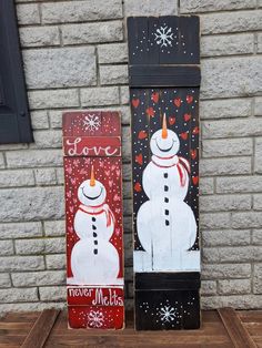 two snowmen painted on wooden boards sitting next to each other in front of a brick wall