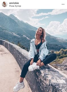 a woman sitting on top of a stone wall next to a lush green forest covered hillside