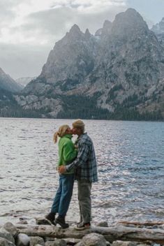 a man and woman standing next to each other on rocks near water with mountains in the background