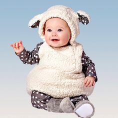 a baby in a sheep costume sitting on the floor with her hands up and smiling