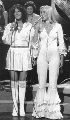 two women in white outfits are singing into microphones while another woman stands behind them