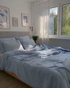 an unmade bed with blue sheets and pillows in front of a large window overlooking the woods