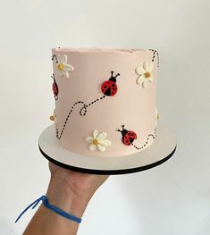 a hand holding a pink cake with ladybugs and daisies on the side