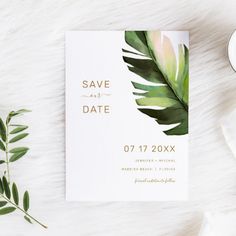 a white save the date card with green leaves on it next to a cup of coffee