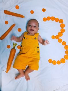 a baby in yellow overalls laying on a bed with carrots scattered around it