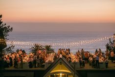 a group of people sitting at tables next to the ocean with lights strung over them