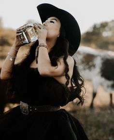 a woman in a black dress and hat drinking water from a bottle while standing next to a cow