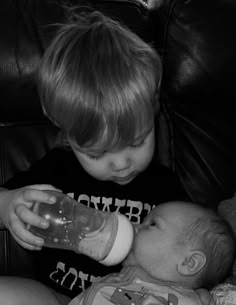 a black and white photo of a young boy drinking from a bottle while sitting next to a baby