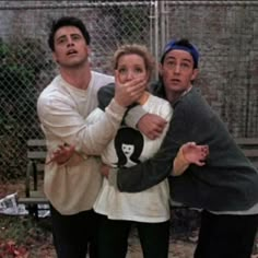 three people standing next to each other in front of a chain link fence with one person covering his mouth