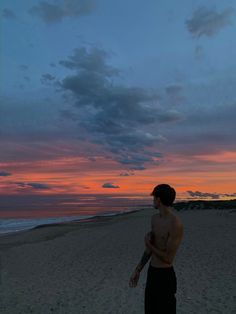 a man standing on top of a sandy beach next to the ocean at sunset with clouds in the sky