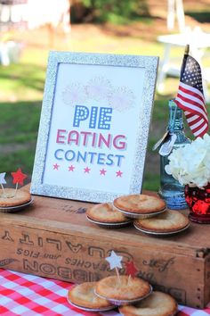 pies are sitting on a picnic table with an american flag in the background and a sign that says pie eating contest