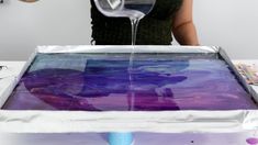 a woman pouring liquid into a pan with purple and blue paint on the bottom, in front of a white wall