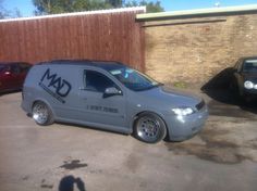 a grey van parked in a parking lot next to two black cars and a wooden fence