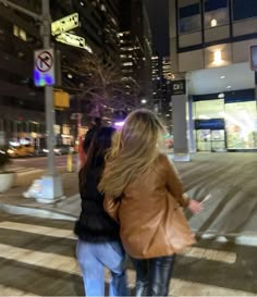 two people walking across a cross walk at night in front of tall buildings and street signs