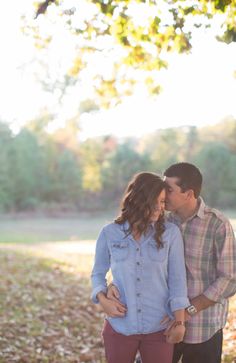 a man and woman standing next to each other under a tree in the fall with leaves on the ground