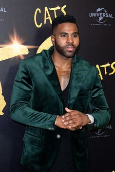 a man in a green jacket and black shirt standing on a red carpet with his hands together