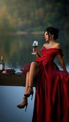 a woman in a red dress sitting on a dock holding a wine glass and bottle