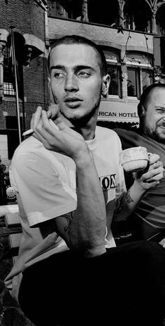 two men sitting next to each other in front of a building and one holding a drink