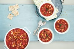 three bowls filled with soup next to crackers and spoons on a blue surface