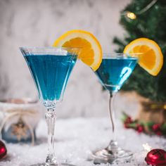 two martini glasses with blue liquid and orange slices on the rims in front of a christmas tree