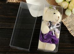 a white teddy bear with a purple ribbon in a clear box on a wooden table