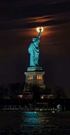 the statue of liberty is lit up at night in new york city, with full moon behind it