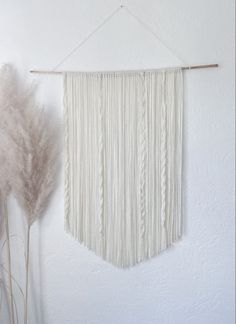 a white wall hanging next to some dried plants and a plant with long stems in front of it