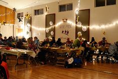 many people are sitting at tables in a room with lights strung from the ceiling and decorations on the walls