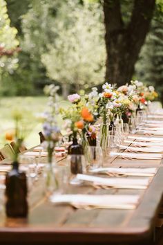 a long table with many vases and glasses on it, set for an outdoor dinner