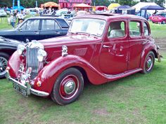 an old red car parked on top of a lush green field next to other cars