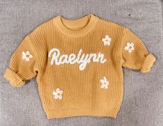 a yellow sweater with white flowers on the chest and name raelynn written on it