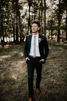 a man wearing a suit and tie standing in the woods with his hands on his hips