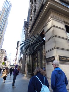 two people walking down the sidewalk in front of a building on a city street with tall buildings behind them