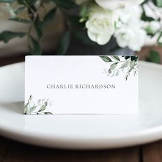 a place card sitting on top of a white plate next to some flowers and greenery