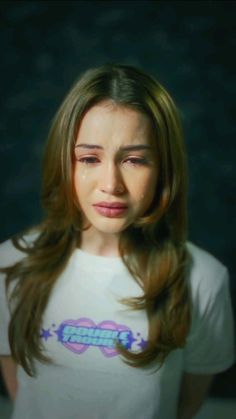 a woman with long brown hair wearing a white t - shirt and looking at the camera