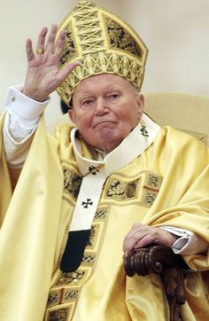 an older man in a priest's outfit waves to the crowd from his chair