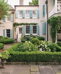 an old house with blue shutters and flowers in the front yard, surrounded by greenery