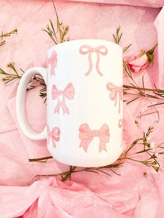 a white coffee mug with pink bows on it and flowers in front of the cup