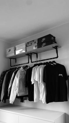 black and white photograph of clothes hanging on rail in room with shelf, drawers and chest