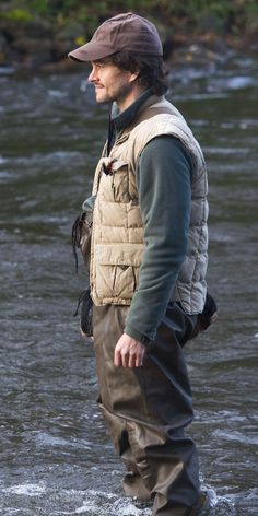 a man standing in the middle of a river wearing a hat and wader pants