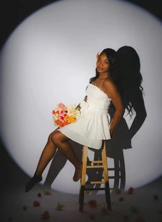 a woman in a white dress sitting on a stool with flowers around her and holding a bouquet