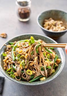 a person holding chopsticks over a bowl of noodles and vegetables with peanuts on the side