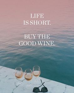 two wine glasses sitting on top of a table next to the ocean with a quote about life is short buy the good wine