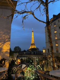 the eiffel tower is lit up at night from an outdoor seating area with tables and chairs