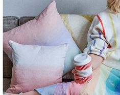 a woman sitting on a couch with a cup of coffee in her hand next to pillows