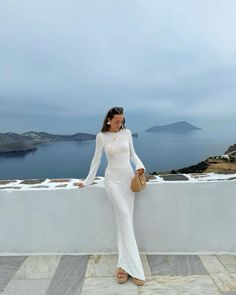 a woman standing on top of a white wall next to the ocean wearing a dress