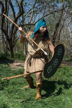 a man dressed as a roman warrior holding a spear and shield in the grass with trees behind him