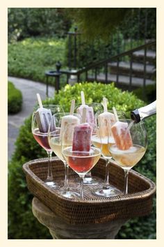 wine glasses filled with different types of drinks on top of a wooden tray in front of bushes