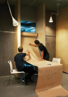 two men working on an unfinished table in a room with large windows and lamps hanging from the ceiling